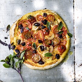 Buchcover „Deftig vegan mediterran“ mit goldbrauner Gemüsequiche, dekoriert mit Tomaten, Oliven und Kräutern.